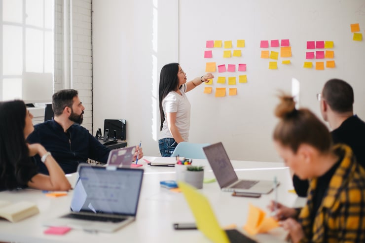 individuals meeting around a table, looking at a women putting colorful stick notes on a board. Photo by Jason Goodman, courtesy of Unsplash