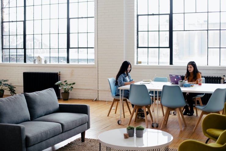 A picture of a room with a couch and coffee table in the foreground, and two women working at a table in the background. Photo by Jason Goodman courtesy of Unsplash